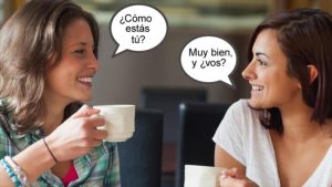 One woman asking ""¿Cómo estás tú? and another woman answering "Muy bien, y ¿vos?" showcasing US Spanish translations